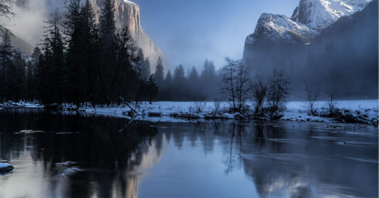Snowy mountains and trees reflecting on water.