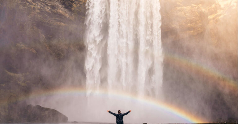 rainbow person water featured image Person standing underneath a waterfall with a rainbow.