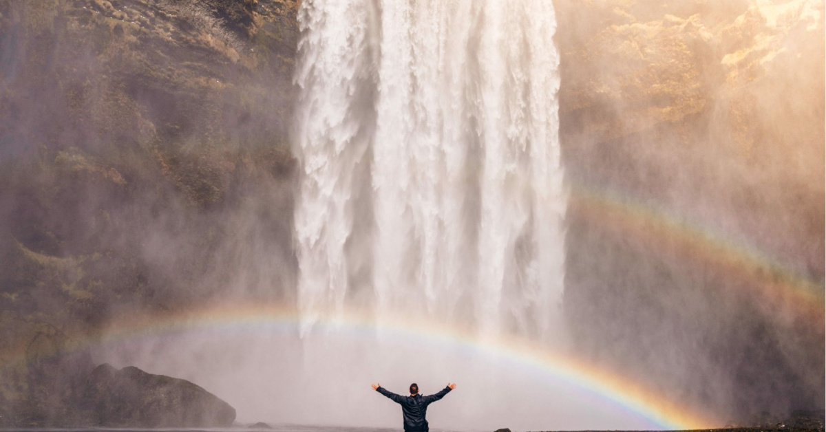 Person standing underneath a waterfall with a rainbow.