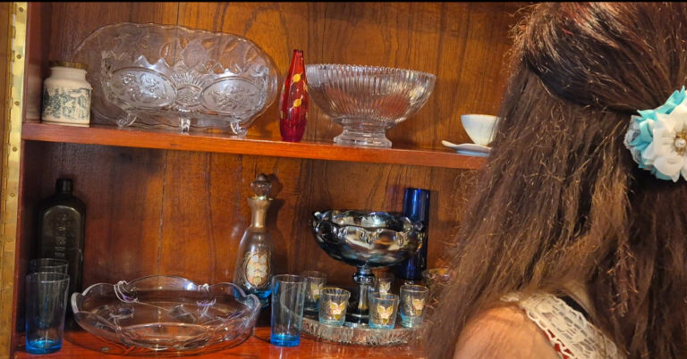 An antique wooden desk, filled with antique glassware and back of Angela Woolcott author looking in.