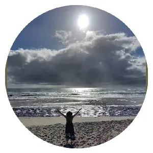 Angela Woolcott, standing on golden sand, facing the morning sun, white clouds, and ocean on Belongil beach, Byron Bay, with arms outstretched. 