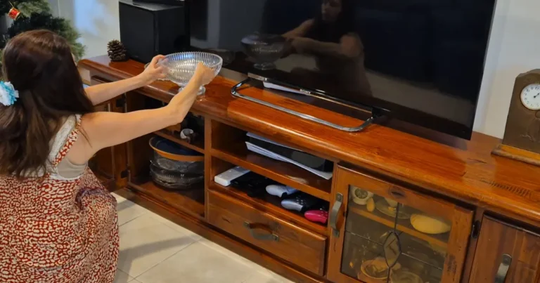 Angela Woolcott, with long dark hair and wearing a patterned dress, kneeling while organising glass bowls and household items within a large wooden entertainment unit, next to a flat-screen television.