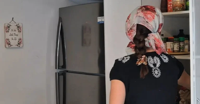 A woman with a long dark braid and a floral headscarf is seen from behind, opening a white kitchen pantry filled with spice jars next to a stainless steel refrigerator.