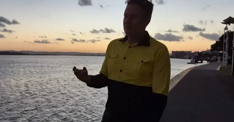 Brad standing on a paved riverfront path at sunset, wearing a yellow and navy high-visibility work shirt and gesturing with his hand as he talks.