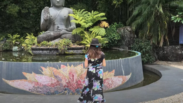 Woman in a floral dress and hat standing in front of a stone Buddha statue and lotus mosaic pond at Crystal Castle in the Byron Shire hinterland