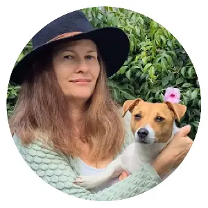 Angela Woolcott holding a small brown and white dog with a pink flower tucked behind its ear, set against a leafy green background.